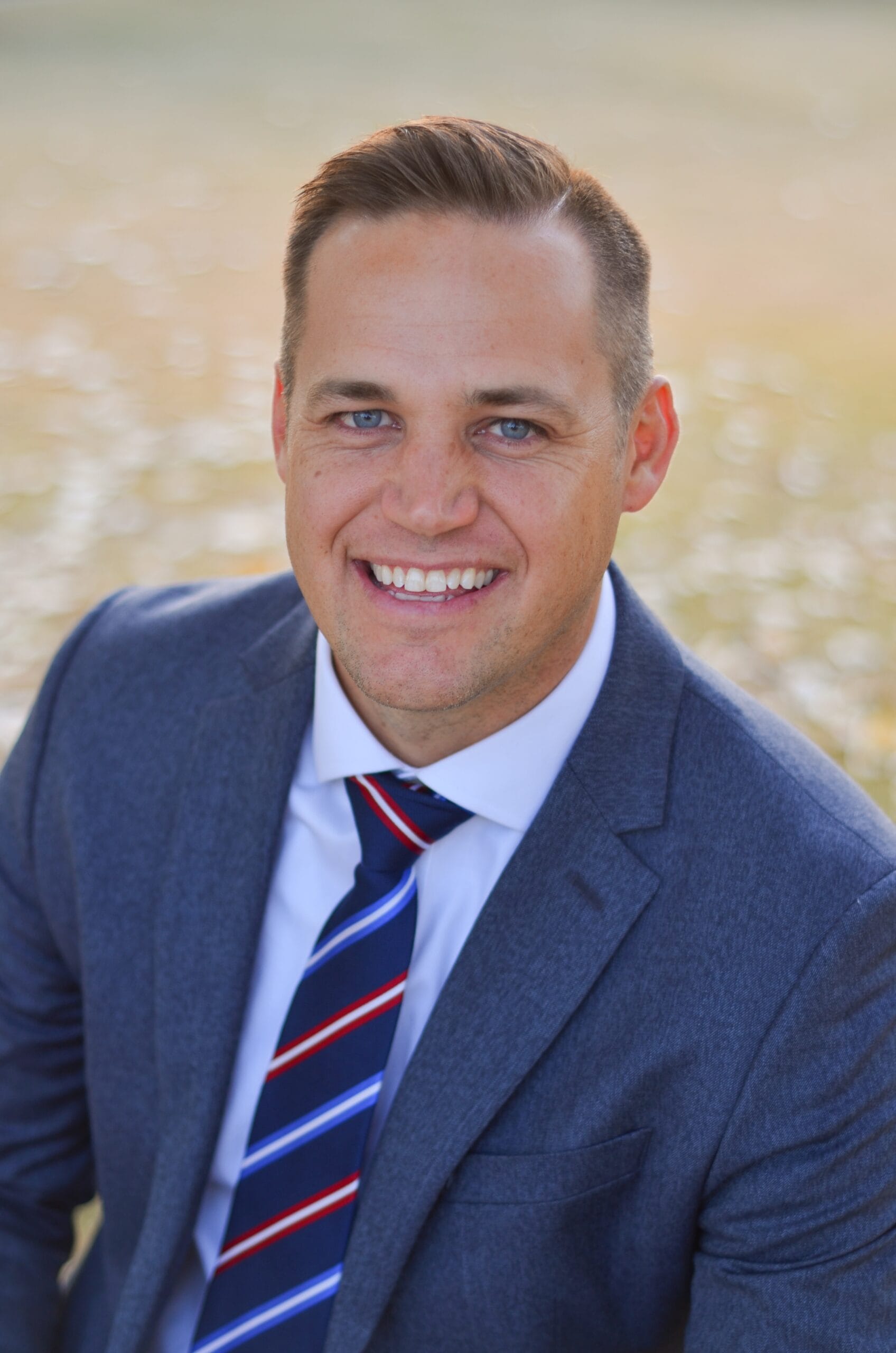 Professional headshot of Ryan Buchanan wearing a blue suit and striped tie, smiling outdoors.